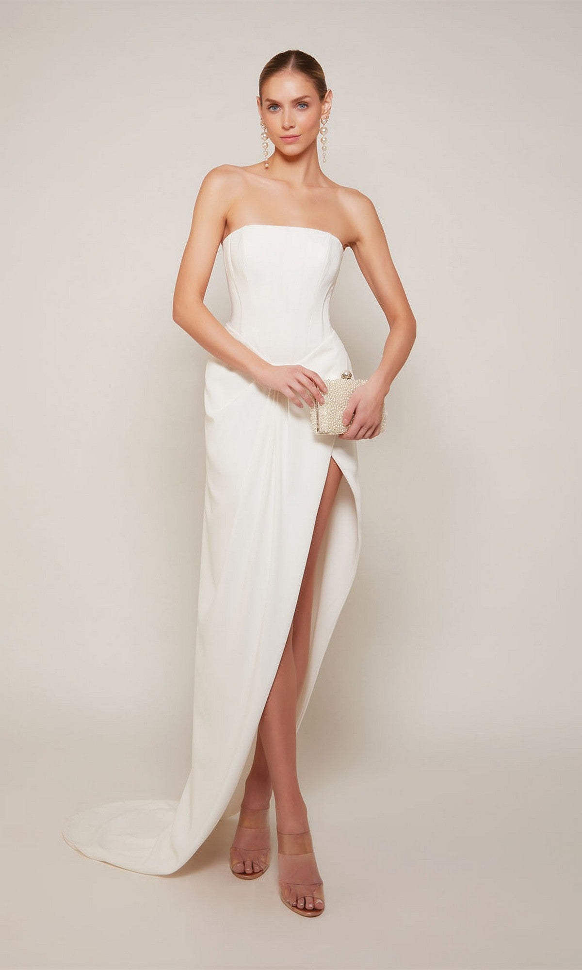 Wearing the Long Formal Dress: Alyce 70042 with a strapless corset bodice and high slit sheath skirt, a woman holds a small clutch and poses against a plain background.